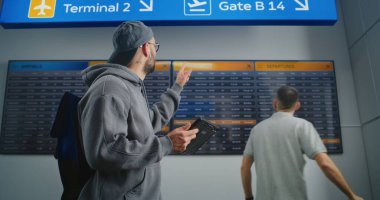 Yoğun Havaalanı Terminali: Sırt çantalı Gezgin Tablet Bilgisayar Kullanır, Dijital Ayrılış Tahtasındaki Uçuş Bilgilerini Denetler, Tatil Gezisinde Gider. Arka plandaki Yolcular için Havaalanı Zaman Çizelgesi Görünümü.