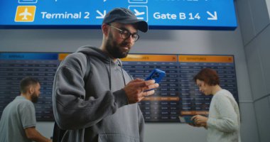 Kalabalık Havaalanı Terminali: Sırt çantalı adam Uçuş Tarayıcı Biletini Telefon kullanarak Kontrol Ediyor, Dijital Uçuş Bilgileri Ekranına Bakıyor. Tatile çıkan bir turist. Arka planda uçaklar kalkış tahtası.