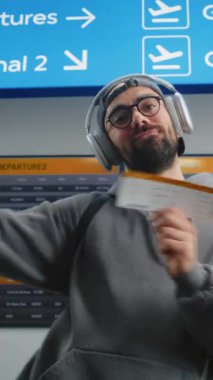 Kalabalık Havaalanı Terminali: Neşeli Dans Eden Adam Portresi, Uçak Bileti Tutma, Kameraya Bakma. Arka planda Dijital Uçuş Bilgileri Ekranı. Gezgin Tatile Çıkıyor. Dikey Çekim.