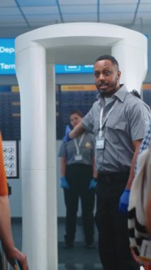 Havaalanı Güvenlik Kontrol Noktası: Afro-Amerikan Güvenlik Memuru, Yolculara Metal Detektör Tarayıcısı, TSA Tarama Prosedürleri Hakkında Bilgi Veriyor. Gezginler Kuyruğu Seyahate Çıkıyor. Dikey Çekim.