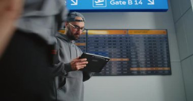 Yoğun Havaalanı Terminali: Sırt çantalı Gezgin Tablet Bilgisayar Kullanır, Dijital Ayrılış Tahtasındaki Uçuş Bilgilerini Denetler, Tatil Gezisinde Gider. Arka plandaki Yolcular için Havaalanı Zaman Çizelgesi Görünümü.