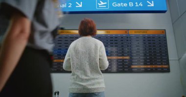 Havaalanı Terminali: Bagajlı Kadın 'ın Arka Görünümü ve Dijital Ayrılış Panosunda Bilet Kontrol Uçuşu. Çeşitli İnsanlar ve Havalimanı Personeli Arka planda Yürüyor. Uçuş Bilgileri Ekranı. Yavaş Hareket