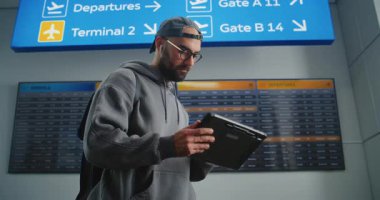 Kalabalık Havaalanı Terminali: Sırt çantalı adam Tablet Bilgisayar kullanıyor, Dijital Uçuş Bilgi Ekranından Gidişini Denetler. Turist Tatile Gidiyor. Arkaplanda Havalimanı Zaman Çizelgesi Görüntüsü.