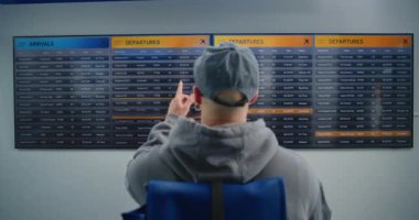 Havaalanı Terminali: Sırt çantalı Adam 'ın Dijital Gidiş Tahtasında Yolcular İçin Uçuşunu Kontrol Etmesi. Gezgin Tatile Çıkıyor. Çeşitli İnsanlar ve Havalimanı Personeli Arka planda Yürüyor.