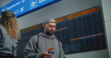 Kalabalık Havaalanı Terminali: Mutlu Adam Uçuş Bilgilerini Kontrol Etmek için Akıllı Telefonu kullanarak Uçak Tarama Biletini Kullanıyor. Arkaplanda Dijital Geliş ve Gidiş Bilgi Ekranı. Gezgin Tatile Çıkıyor.