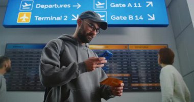 Kalabalık Havaalanı Terminali: Sırt çantalı adam Uçuş Tarayıcı Biletini Telefon kullanarak Kontrol Ediyor, Dijital Uçuş Bilgileri Ekranına Bakıyor. Tatile çıkan bir turist. Arka planda uçaklar kalkış tahtası.