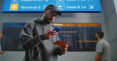 Yoğun Havaalanı Terminali: Sırt çantalı adam Uçuş Bilgi Tarama Biletini Telefon Kullanarak Kontrol Ediyor. Gezgin Tatile Çıkıyor. Arka plandaki Yolcular için Dijital Geliş ve Gidiş Kurulu.