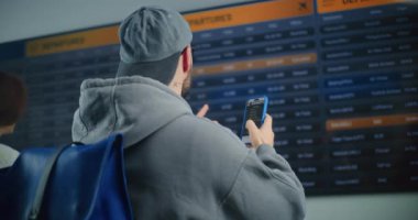 Kalabalık Havaalanı Terminali: Gelen ve Giden Uçuş Bilgilerini Arayan Sırt çantalı Adam, Biniş Uçağı Bilgilerini Kontrol Etmek için Akıllı Telefon 'u kullanıyor. Turist Tatile Çıkıyor.