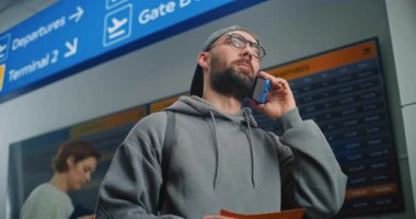 Havaalanı Terminali: Erkek Yolcu Tutma Uçak Bileti, Uçuş Bilgilerini Kontrol Etmek için Telefonla Konuşuyor. Arkaplanda İnsanlar ve Dijital Geliş ve Gidiş Bilgi Ekranı. Turist Seyahate Çıktı.