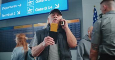 Kalabalık Havaalanı Terminali: Uçağı tutan yaşlı adam, uçuş bilgilerini kontrol etmek için cep telefonuyla konuşuyor. Arka planda, Dijital Geliş ve Gidiş Kurulu, Çeşitli İnsanlar Yürüyor.