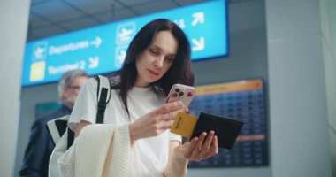 Kalabalık Havaalanı Terminali: Yetişkin Kadın Uçuş Bilgilerini Kontrol Etmek için Telefon Kullanarak Uçak Tarama Biletini Kullanıyor. Arka planda, Dijital Geliş ve Gidiş Kurulu, Çeşitli İnsanlar Yürüyor. Gezgin Seyahate Çıkıyor.