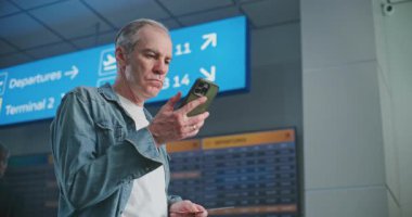 Kalabalık Havaalanı Terminali: Olgun Adam Uçak Tarama Biletini Uçuş Bilgilerini Kontrol Etmek için Telefon Kullanıyor. Gezgin Seyahate Çıkıyor. Arka planda, Dijital Geliş ve Gidiş Görünümü, Çeşitli İnsanlar Yürüyor