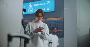 Kalabalık Havaalanı Terminali: Genç adam cep telefonunda kayıyor, biniş için bekliyor. Gezgin Seyahate Çıkıyor. Arka planda, Dijital Geliş ve Gidiş Görünümü, Yürüyen Çeşitli İnsanlar.