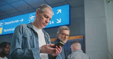 Kalabalık Havaalanı Terminali: Olgun Adam Uçak Tarama Biletini Uçuş Bilgilerini Kontrol Etmek için Telefon Kullanıyor. Gezgin Seyahate Çıkıyor. Arka planda, Dijital Geliş ve Gidiş Görünümü, Çeşitli İnsanlar Yürüyor