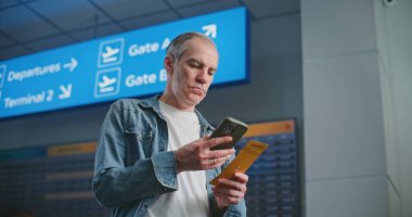 Kalabalık Havaalanı Terminali: Olgun Adam Uçak Tarama Biletini Uçuş Bilgilerini Kontrol Etmek için Telefon Kullanıyor. Gezgin Seyahate Çıkıyor. Arka planda, Dijital Geliş ve Gidiş Görünümü, Çeşitli İnsanlar Yürüyor