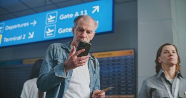 Kalabalık Havaalanı Terminali: Olgun Adam Uçak Tarama Biletini Uçuş Bilgilerini Kontrol Etmek için Telefon Kullanıyor. Gezgin Seyahate Çıkıyor. Arka planda, Dijital Geliş ve Gidiş Görünümü, Çeşitli İnsanlar Yürüyor