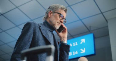 Kalabalık Uluslararası Havaalanı Terminali: Olgun Adam Uçak Tutma Biletleri, Uçuş Bilgilerini Kontrol Etmek için Cep Telefonu ile Konuşma. Gezgin Tatile Çıkıyor. Arka planda Yürüyen Farklı İnsanlar.