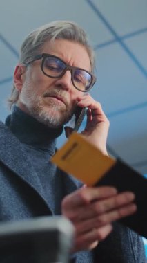 Kalabalık Uluslararası Havaalanı Terminali: Olgun Adam, İş Adamı Tutucu Uçak Bileti, Uçuş Bilgilerini Kontrol Etmek için Cep Telefonu ile Konuşma. Seyahate çıkan gezgin ya da iş gezisi. Dikey Çekim.