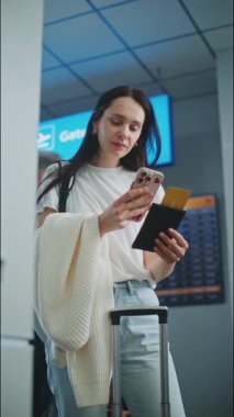Havaalanı Terminali: Yetişkin Kadın Uçuş Bilgilerini Kontrol Etmek için Akıllı Telefon kullanarak Uçak Tarayıcı Biletleri. Dijital Geliş ve Gidiş Ekranı, Farklı İnsanlar Yürüyor. Seyahate çıkan bir turist. Dikey Çekim.