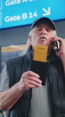 Kalabalık Havaalanı Terminali: Uçağı tutan yaşlı adam, uçuş bilgilerini kontrol etmek için cep telefonuyla konuşuyor. Arka planda, Geliş ve Gidiş Kurulu, Çeşitli İnsanlar Yürüyor. Dikey Çekim.