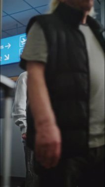 Kalabalık Havaalanı Terminali: Cep Telefonuyla Konuşan Genç Adam Video Çağrısı veya Video Kaydı İçeriği. Geliş ve Gidiş Ekranı, Farklı İnsanlar Yürüyor. Seyahate çıkan bir turist. Dikey Çekim.