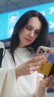Kalabalık Havaalanı Terminali: Yetişkin Kadın Uçuş Bilgilerini Kontrol Etmek için Telefon Kullanarak Uçak Tarama Biletini Kullanıyor. Dijital Geliş ve Gidiş Kurulu, Çeşitli İnsanlar Yürüyor. Gezgin Seyahate Çıkıyor. Dikey Çekim.