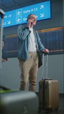 Uluslararası Havaalanı Terminali: Olgun Adam Bavulla Duruyor, Uçuş Bilgilerini Kontrol Etmek İçin Cep Telefonu ile Konuşuyor. Dijital Geliş ve Gidiş Görünümü, Farklı İnsanlar Yürüyüşü. Dikey Çekim.