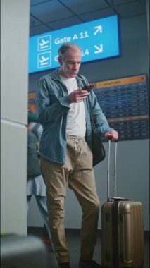 Kalabalık Havalimanı Terminali: Akıllı Telefondan Bavul Kaydıran Olgun Adam, Uçuş Bekliyor. Gezgin Seyahate Çıkıyor. Geliş ve Gidiş Görünümü, Farklı İnsanlar Yürüyor. Dikey Çekim.