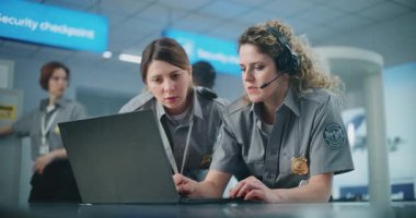 Havaalanı Güvenlik Kontrol Noktası: Kulaklık takan kadın güvenlik görevlisi, iş arkadaşı ile birlikte laptop kullanıyor. Seyahat Güvenliği Ulaştırma Güvenlik İdaresi. Arka planda, havalimanı personeli çalışıyor.