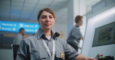 Havaalanı Güvenlik Kontrol Noktası: Metal dedektör kontrol panelinde oturan ve kameraya bakan kadın TSA memurunun portresi. Gelişmiş bagaj tarama prosedürünün X-ray görüntülerini gösteren bilgisayar monitörleri.