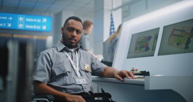Havaalanı Güvenlik Kontrol Noktası: Kameraya bakan Afrikalı Amerikalı TSA işçisinin portresi. Bilgisayar ekranları bagaj tarama prosedürünün X-ray görüntülerini gösteriyor. Arka planda, havaalanı personeli çalışıyor.