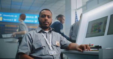 Havaalanı Güvenlik Kontrol Noktası: Kameraya bakan Afrikalı Amerikalı TSA işçisinin portresi. Bilgisayar ekranları bagaj tarama prosedürünün X-ray görüntülerini gösteriyor. Arka planda, havaalanı personeli çalışıyor.