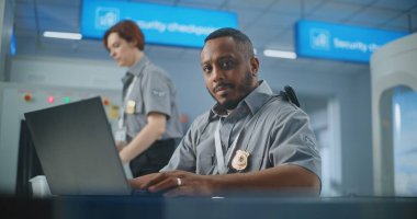 Havaalanı Güvenlik Kontrol Noktası: Afro-Amerikan Güvenlik Subayı Laptop Bilgisayar kullanıyor, Kameraya bakıyor. Seyahat Güvenliği Ulaştırma Güvenlik İdaresi. Arka planda, havalimanı personeli çalışıyor.