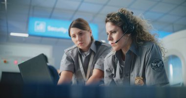 Havaalanı Güvenlik Kontrol Noktası: Kulaklık takan kadın TSA memuru Laptop kullanıyor, İş arkadaşı ile konuşuyor. Seyahat Güvenliği Ulaştırma Güvenlik İdaresi. Arka planda, havaalanı personeli çalışıyor.