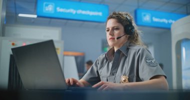 Havaalanı Güvenlik Kontrol Noktası: Laptop 'ta kulaklık takan kadın TSA memuru. Seyahat Güvenliği Ulaştırma Güvenlik İdaresi. Arka planda, havalimanı personeli tarama prosedürleri.