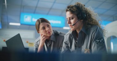 Havaalanı Güvenlik Kontrol Noktası: Kulaklıklı iki kadın güvenlik görevlisi dizüstü bilgisayar kullanarak konuşuyor. Seyahat Güvenliği Ulaştırma Güvenlik İdaresi. Arka planda, havaalanı personeli çalışıyor.