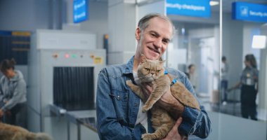 Havaalanı Terminali: Ellerinde Kedi Tutan, Gülümseyen ve Kameraya Bakan Olgun Adamın Portresi. Arka planda, Uçuş Güvenliği Kontrol Noktasında Yolcu ve Evcil Hayvanlar için TSA Tarama Prosedürleri