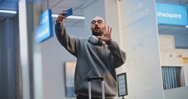 Kalabalık Havaalanı Terminali: Video Call ya da Recording Video Content tarafından telefonla konuşan bavullu turist. Gezgin Tatile Çıkıyor. Arka planda, Çeşitli İnsanlar ve Havaalanı Personeli Yürüyüşü.