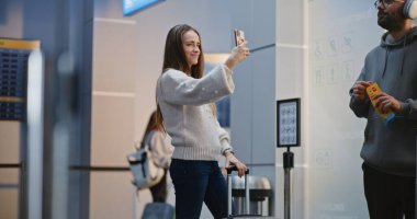 Kalabalık Havalimanı Terminali: Yetişkin Kadın Smartphone 'da Selfie Çekiyor, Havayolu İnbotu Biniş Salonunda Uçuşu Bekliyor, Adamla Konuşuyor. Seyahate çıkan bir turist. Çeşitli İnsanlar ve Havalimanı Personeli Yürüyüşü