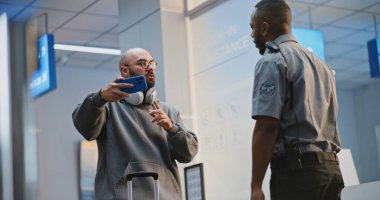 Kalabalık Havaalanı Terminali: Smartphone 'da bavullu bir adam video kayıt yapıyor. Afrikalı Amerikalı güvenlik görevlisi video kaydının yasaklanması konusunda uyardı. Arka planda, Yürüyen Çeşitli İnsanlar.