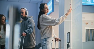 Kalabalık Uluslararası Havaalanı Terminali: Güzel Kadın Smartphone 'da Selfie Çekiyor, Uçağa Bekleme Salonunda Bekliyor. Seyahate çıkan bir turist. Çeşitli İnsanlar ve Havalimanı Personeli Yürüyüşü.