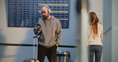 Yoğun Uluslararası Havaalanı Terminali: Kulaklıklı Adam Telefon Kullanıyor, Selfie Çekiyor, Havayolu Hub Salonunda Uçuş Bekliyor. Gezgin Seyahate Çıkıyor. Çeşitli İnsanlar ve Havalimanı Personeli Yürüyüşü.