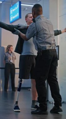 Havaalanı Terminalindeki güvenlik kontrol noktası: Afrikalı Amerikan Güvenlik Subayı Biyonik Protez Bacaklı Genç Adamı Uçuş için Denetliyor. TSA tarama prosedürleri. Engelli turist. Dikey Çekim.