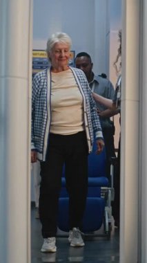 Güvenlik Kontrol Noktasında Kıdemli Gezgin: Tekerlekli sandalyeden Kalkan Yaşlı Kadın, Havaalanı Terminalinde Metal Detektör Tarayıcısından Geçen, Havaalanı Personel Yardımcıları Kadın. Tarama prosedürleri. Dikey Çekim.