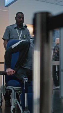 Havaalanı Terminalindeki güvenlik kontrol noktası: Tekerlekli sandalyeden kalkan protez bacaklı adam, X-ray tarayıcısını geçiyor, güvenlik personeli yardım ediyor. Tarama sırasında yolcular. Dikey Çekim.