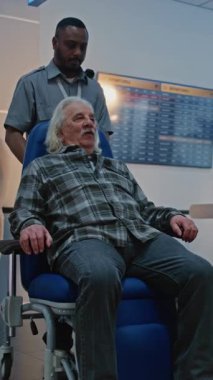 Havaalanındaki güvenlik kontrol noktası: Tekerlekli sandalyeden kalkan yaşlı adam, metal dedektörü tarama kapısından geçiyor, güvenlik görevlisi yardım ediyor. Gösterim sırasında yaşlı turist. Dikey Çekim.