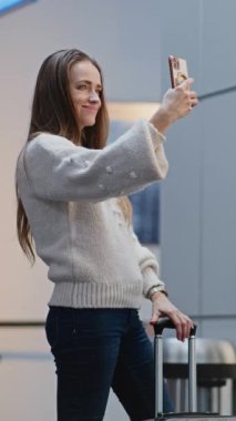 Kalabalık Havaalanı Terminali: Smartphone 'da Selfie Çeken Kadın, Uçağa Binme Merkezi' nde Uçuşu Bekliyor, Konuşuyor. Seyahate çıkan bir turist. Çeşitli insanlar, havaalanı personeli yürüyüşü. Dikey Çekim.