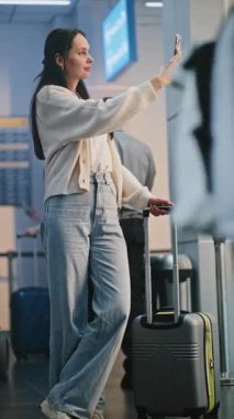 Kalabalık Uluslararası Havaalanı Terminali: Güzel Kadın Smartphone 'da Selfie Çekiyor, Uçağa Bekleme Salonunda Bekliyor. Çeşitli İnsanlar ve Havalimanı Personeli Yürüyüşü. Dikey Çekim.