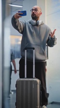 Kalabalık Havaalanı Terminali: Video Call ya da Recording Video Content tarafından telefonla konuşan bavullu turist. Gezgin Tatile Çıkıyor. Çeşitli İnsanlar ve Havalimanı Personeli Yürüyüşü. Dikey Çekim.