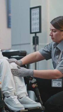 Havaalanı Terminali 'ndeki güvenlik kontrol noktası, kadın güvenlik görevlisi tekerlekli sandalyedeki kadını teftiş etti. TSA tarama prosedürleri. Yolcu ile Yürüyen İşçi Engelli Uçuşu. Dikey Çekim.